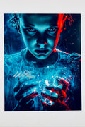 A close-up promotional photo of Millie Bobby Brown as Eleven from Stranger Things, lit in dramatic blue and red tones with glowing energy effects around her hands, bearing a silver signature on the left side.