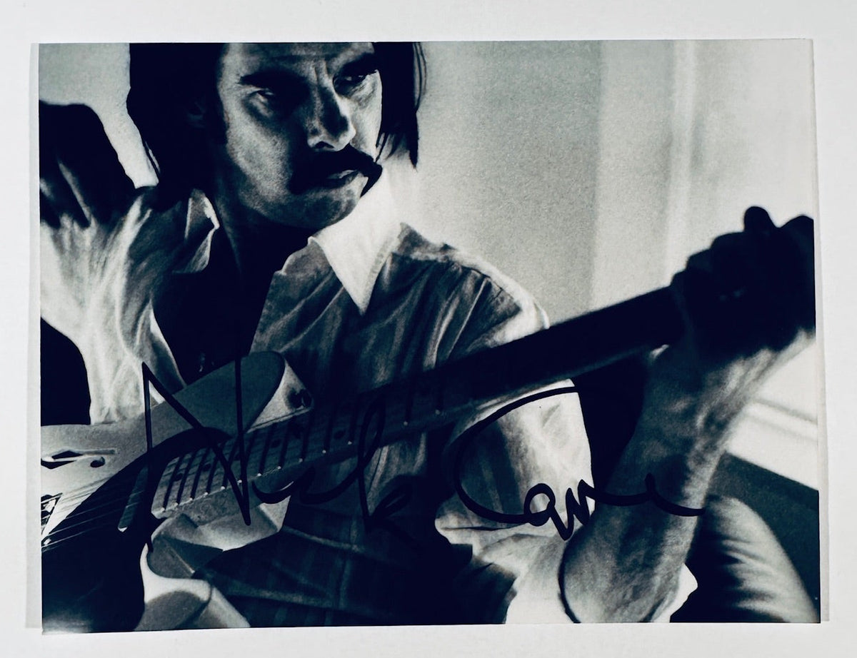 Full landscape layout of a black and white portrait photograph showing musician Nick Cave signed boldly in black ink, isolated against a plain white background.