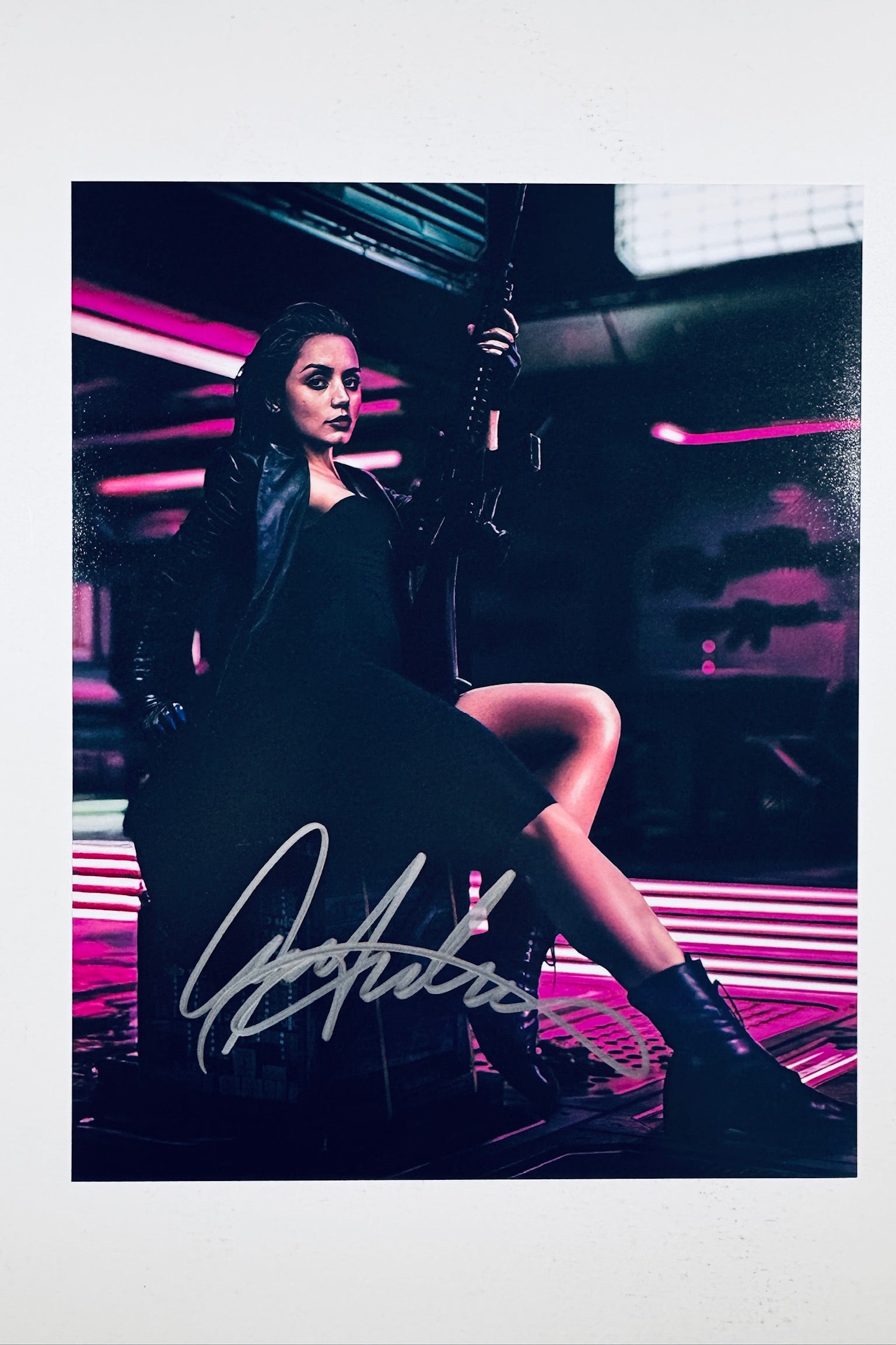 A portrait of Ana de Armas holding a rifle while sitting in a dimly lit cyberpunk setting illuminated by neon pink lights, featuring a silver marker signature across the lower center.