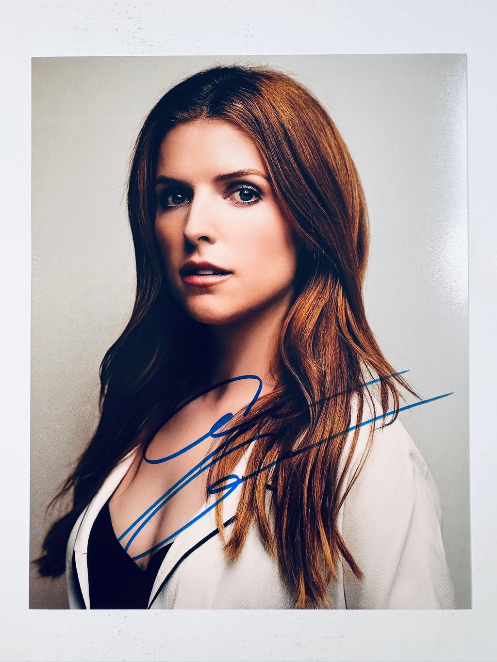 Signed 8x10 matte promotional portrait of actress Anna Kendrick with long brown hair wearing a white jacket and dark top against a light background, featuring a bold blue ink autograph.