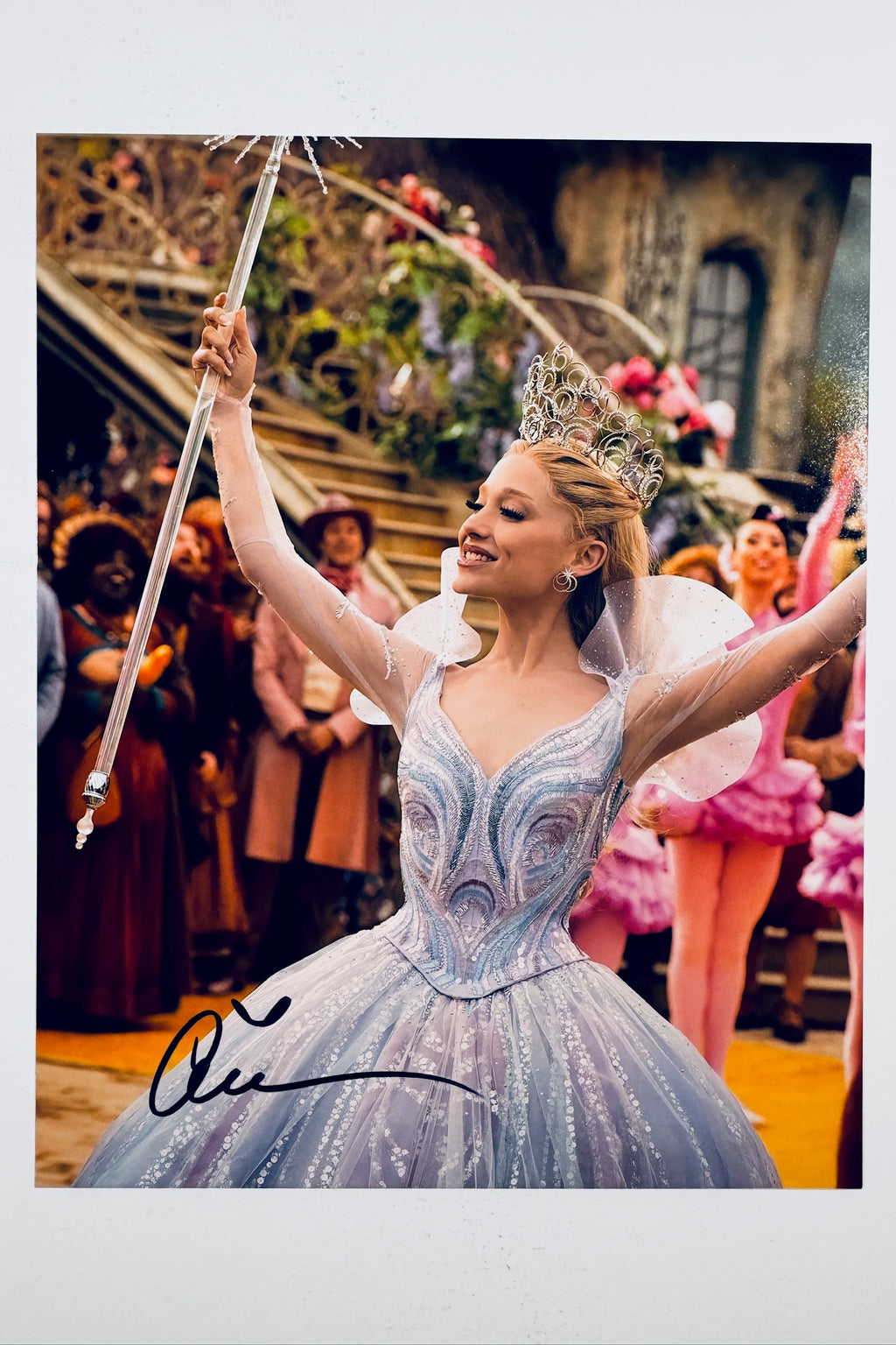 A portrait of Ariana Grande as Glinda in Wicked wearing an ornate crown and light blue gown while holding a wand up high, featuring a black marker signature across the lower left of her dress.