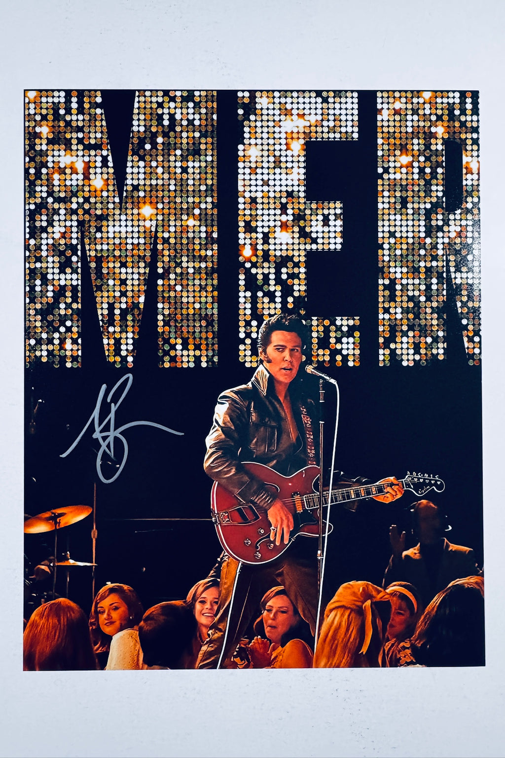 A portrait of Austin Butler as Elvis performing on stage with a red guitar under stage lights and a large sequined backdrop, featuring a silver marker signature on the left.