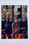 A portrait of Austin Butler as Elvis performing on stage with a red guitar under stage lights and a large sequined backdrop, featuring a silver marker signature on the left.