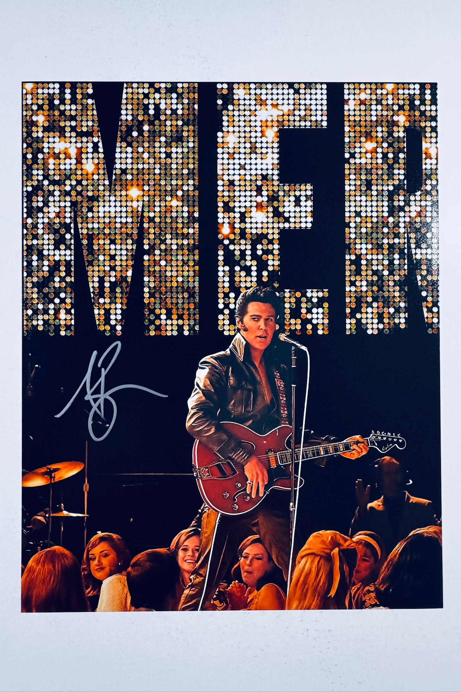 A portrait of Austin Butler as Elvis performing on stage with a red guitar under stage lights and a large sequined backdrop, featuring a silver marker signature on the left.