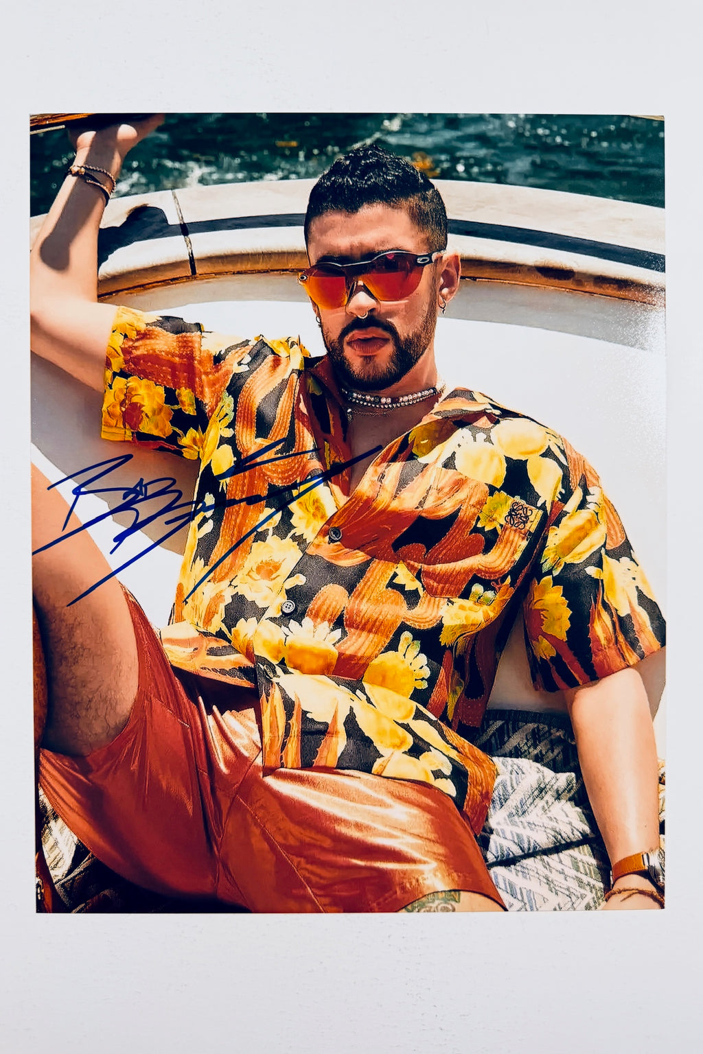 A portrait of Bad Bunny relaxing on a boat wearing a vibrant floral short-sleeve shirt, orange shorts, and tinted sunglasses, featuring a blue marker signature across the left side.