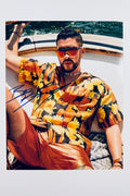 A portrait of Bad Bunny relaxing on a boat wearing a vibrant floral short-sleeve shirt, orange shorts, and tinted sunglasses, featuring a blue marker signature across the left side.