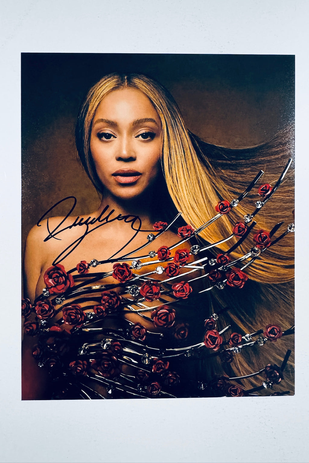A portrait of Beyoncé with blonde and brown hair against a dark background, wearing a strapless outfit adorned with silver branches and red roses, featuring a dark marker signature on the left.