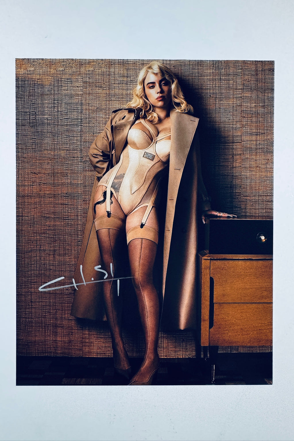 A portrait of Billie Eilish with blonde hair wearing a beige corset-style bodysuit and a tan trench coat, standing against a textured wall, with a white autograph on the lower left.