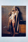 A portrait of Billie Eilish with blonde hair wearing a beige corset-style bodysuit and a tan trench coat, standing against a textured wall, with a white autograph on the lower left.