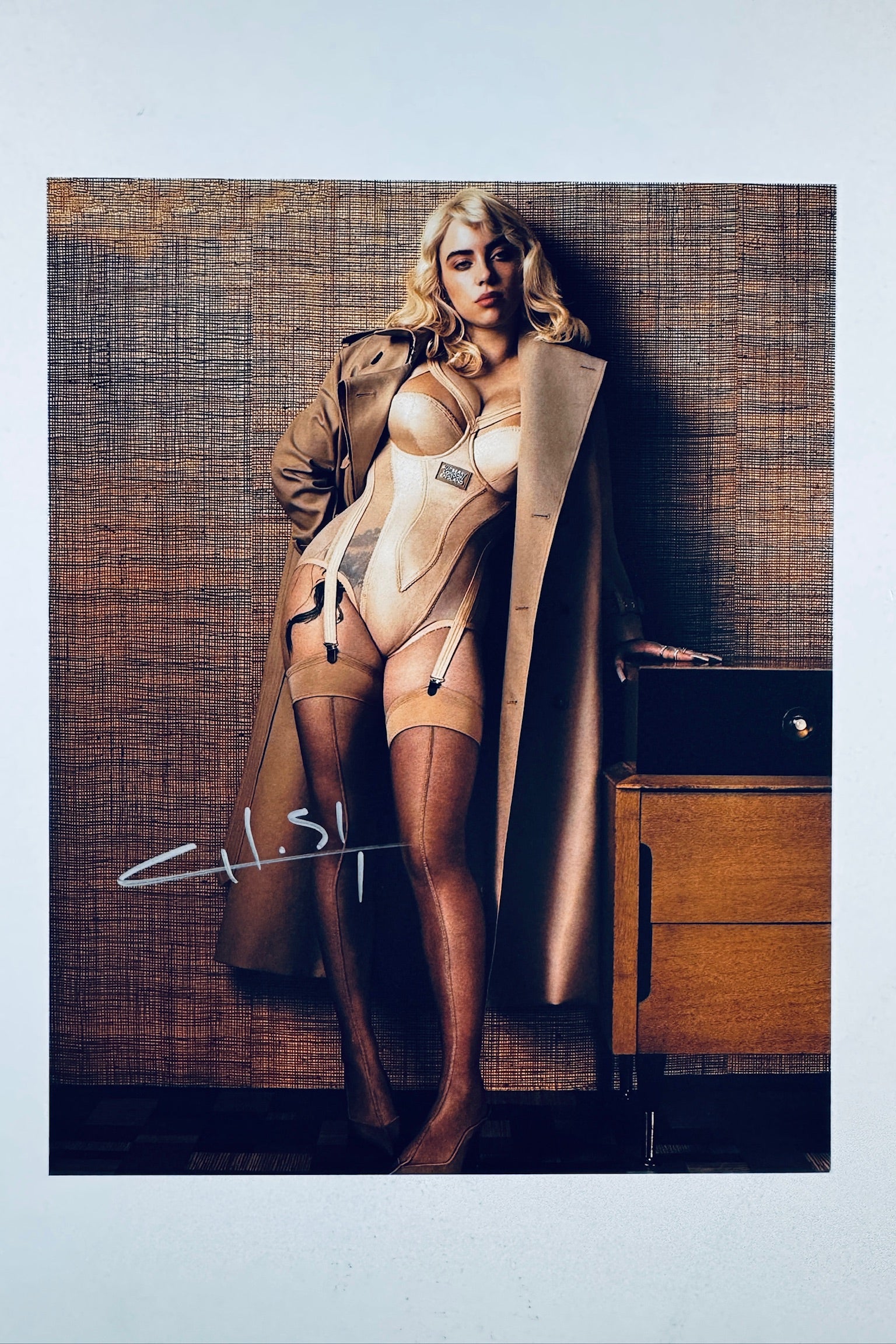A portrait of Billie Eilish with blonde hair wearing a beige corset-style bodysuit and a tan trench coat, standing against a textured wall, with a white autograph on the lower left.
