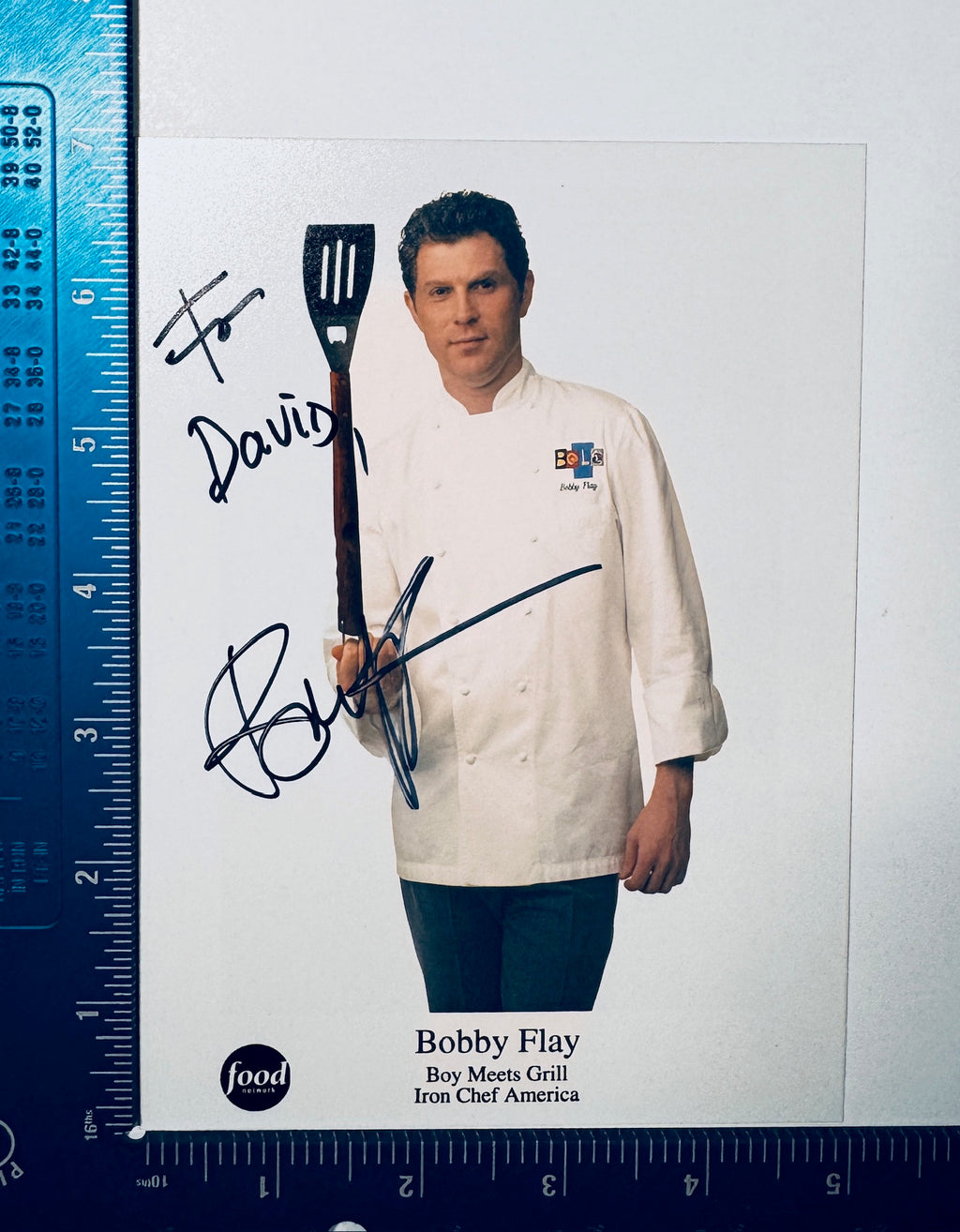 Autographed Bobby Flay signed photo holding a spatula next to a measuring ruler.