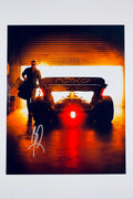 A still from the F1 movie featuring Brad Pitt carrying a bag while walking past an APXGP race car in a garage with dramatic warm lighting, signed in silver ink on the lower left.
