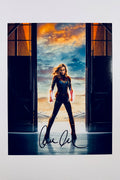 A portrait of Brie Larson as Captain Marvel standing centered in a hangar doorway with glowing fists raised against a cloudy sunset sky, featuring a bold black marker signature across the lower foreground.