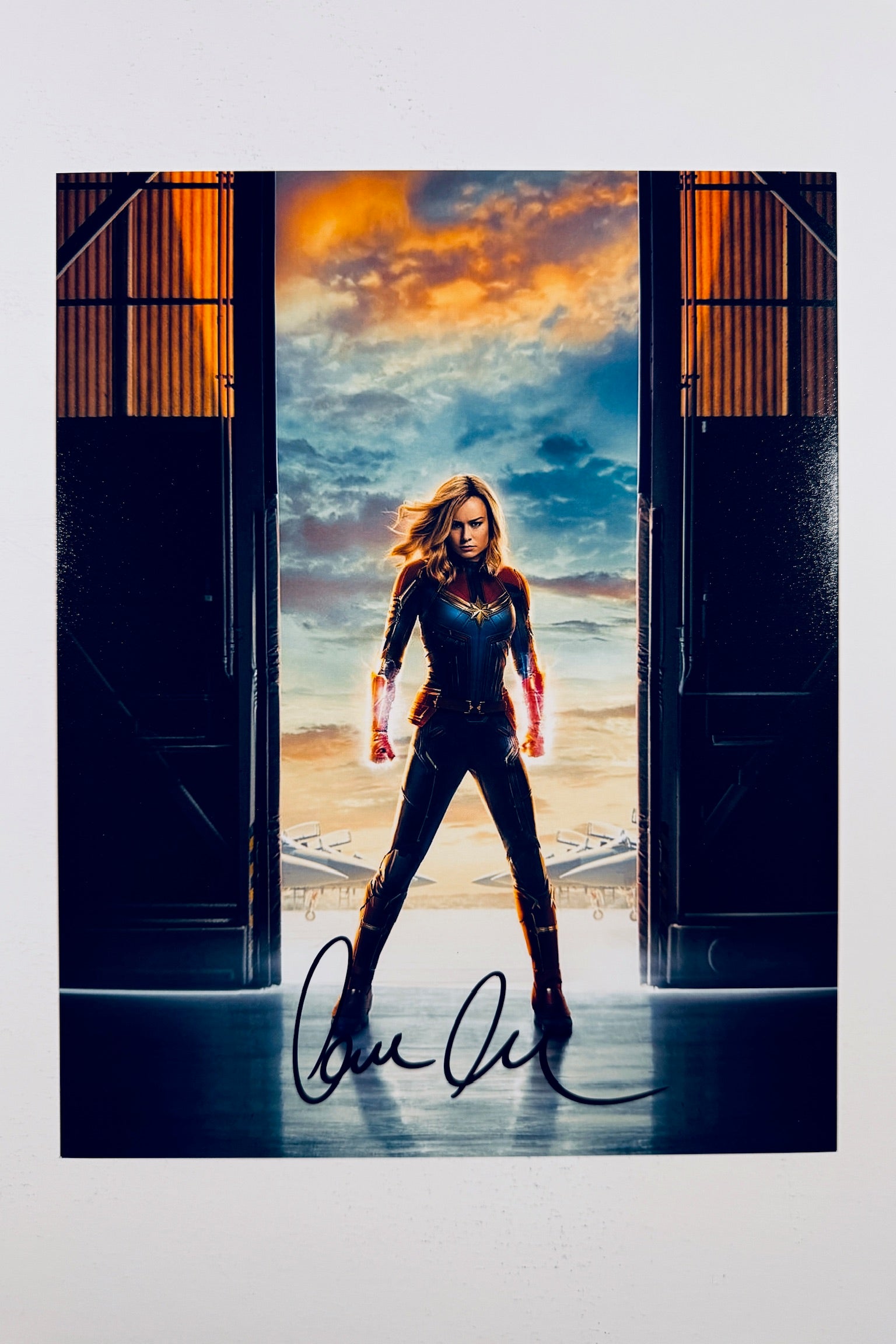 A portrait of Brie Larson as Captain Marvel standing centered in a hangar doorway with glowing fists raised against a cloudy sunset sky, featuring a bold black marker signature across the lower foreground.