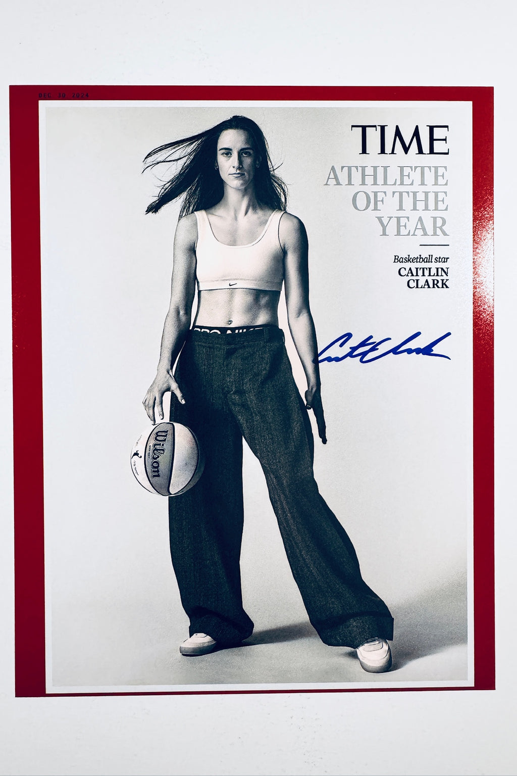 A portrait of basketball star Caitlin Clark styled for Time Athlete of the Year holding a basketball with wide-leg pants and a white sports bra, featuring a blue marker signature on the right.