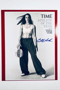 A portrait of basketball star Caitlin Clark styled for Time Athlete of the Year holding a basketball with wide-leg pants and a white sports bra, featuring a blue marker signature on the right.