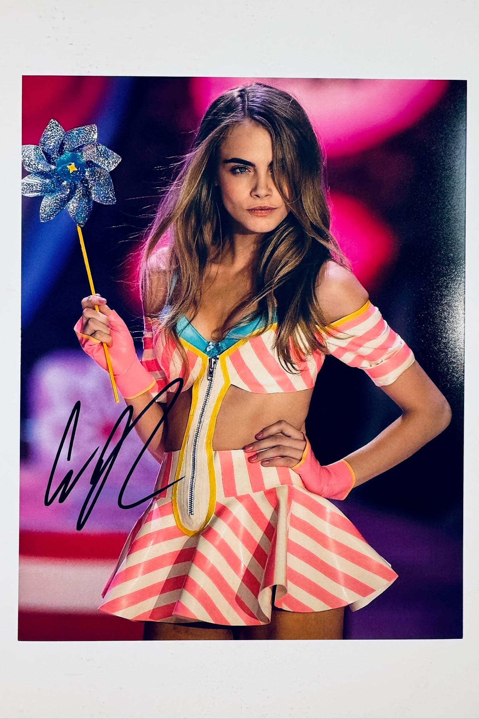 Cara Delevingne hand-signed 8x10 matte photo holding a blue pinwheel on the runway wearing a pink and white striped outfit.