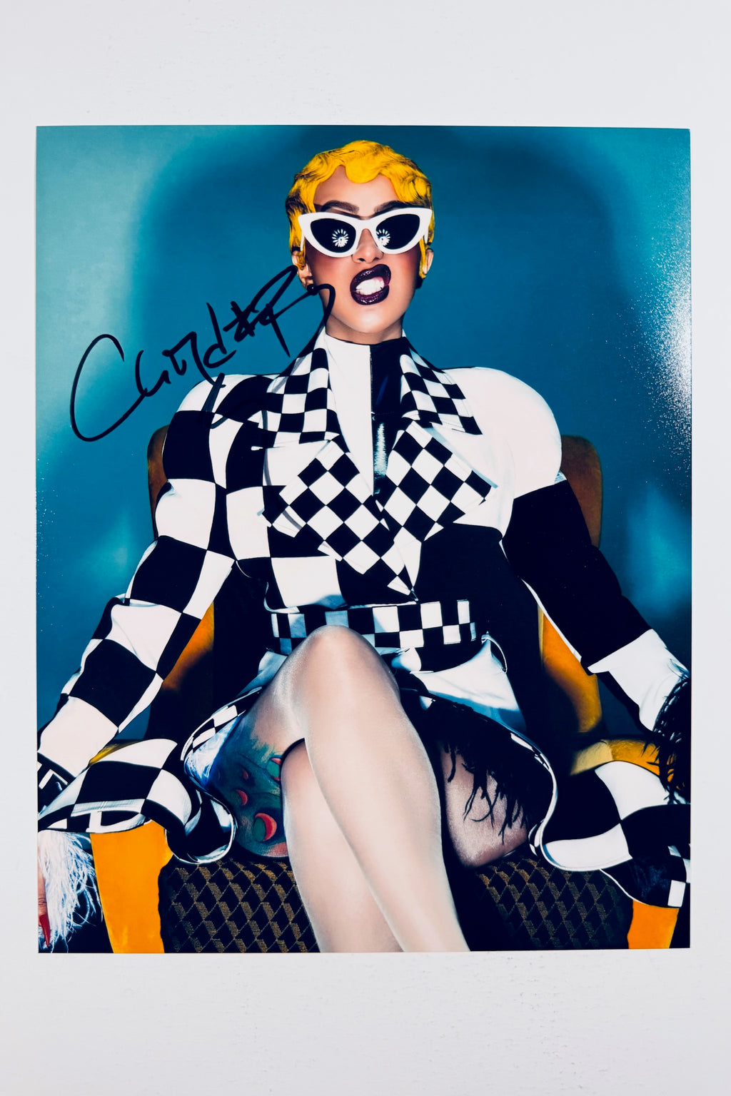 A portrait of Cardi B sitting in a yellow chair wearing a black and white checkerboard jacket, white cat-eye sunglasses, and yellow hair, featuring a dark marker signature on the upper left.