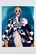 A portrait of Cardi B sitting in a yellow chair wearing a black and white checkerboard jacket, white cat-eye sunglasses, and yellow hair, featuring a dark marker signature on the upper left.