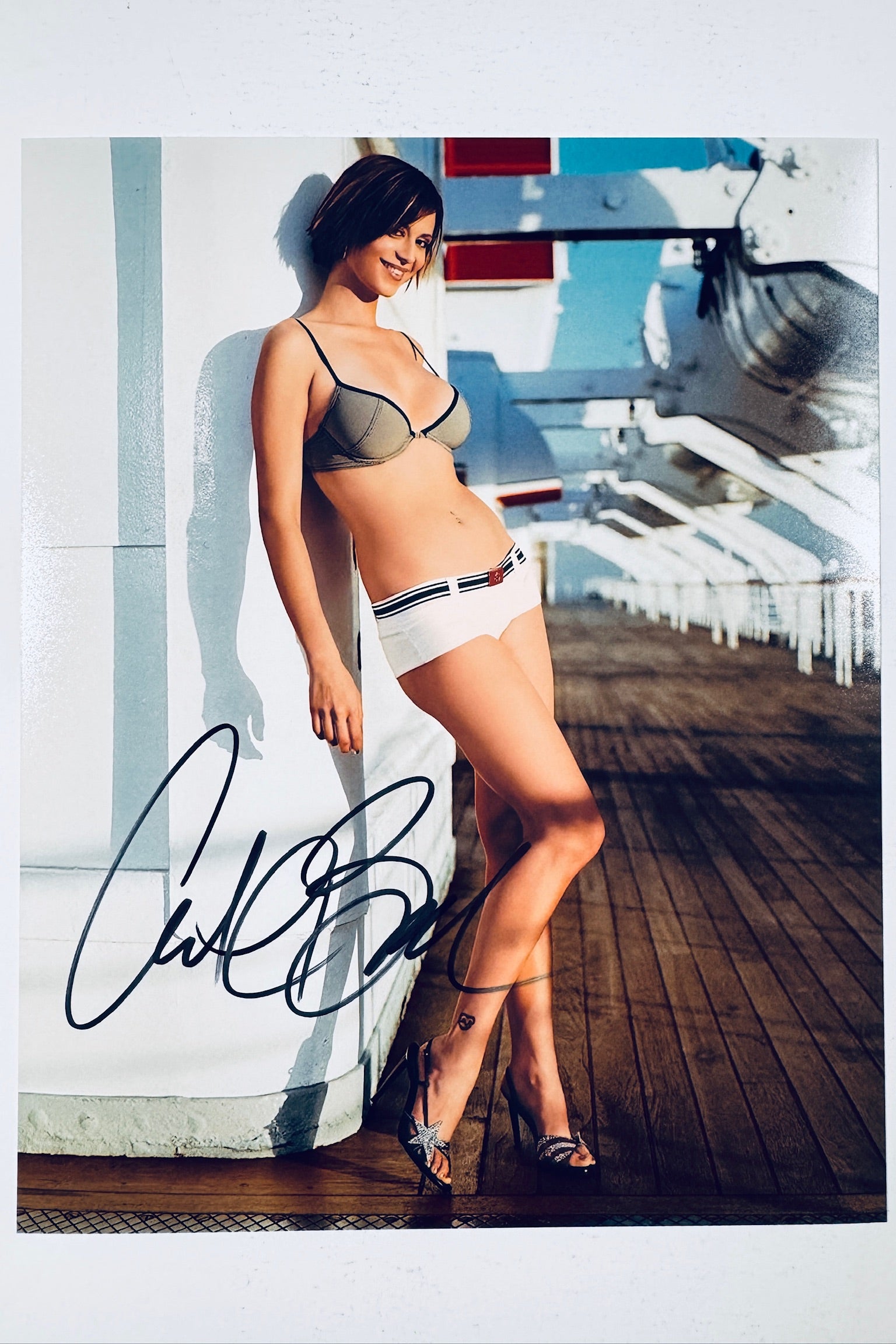 Signed full photo of Catherine Bell leaning against a white structure on a wooden deck wearing a bra and shorts, featuring a black autograph.