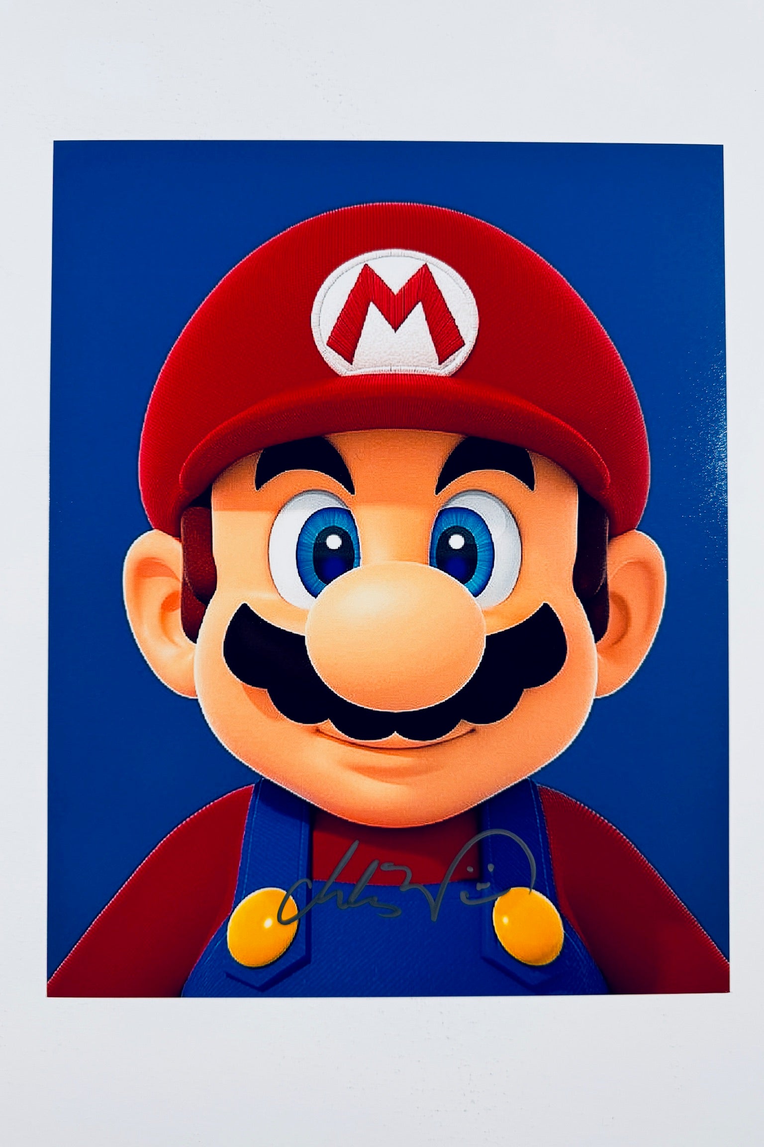 A close-up portrait of Mario with his red hat and blue overalls against a solid blue background, featuring a dark marker signature across his chest straps.