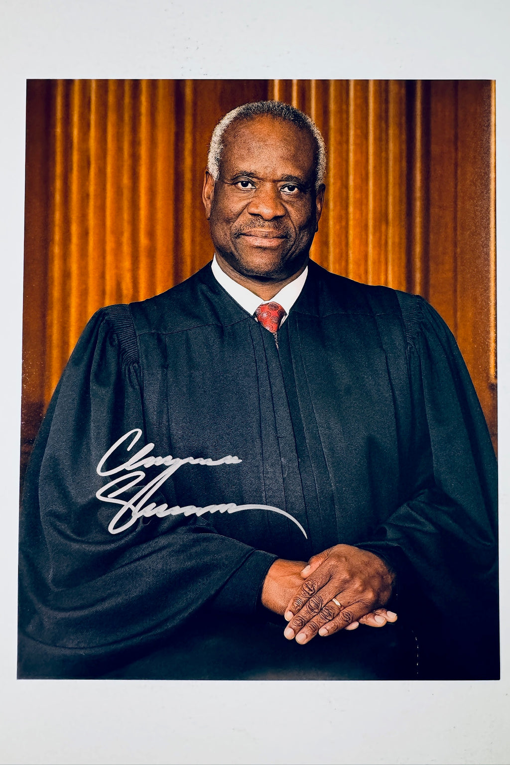 Clarence Thomas hand-signed 8x10 matte photo wearing his judicial robe and patterned tie in front of a wood-paneled background.