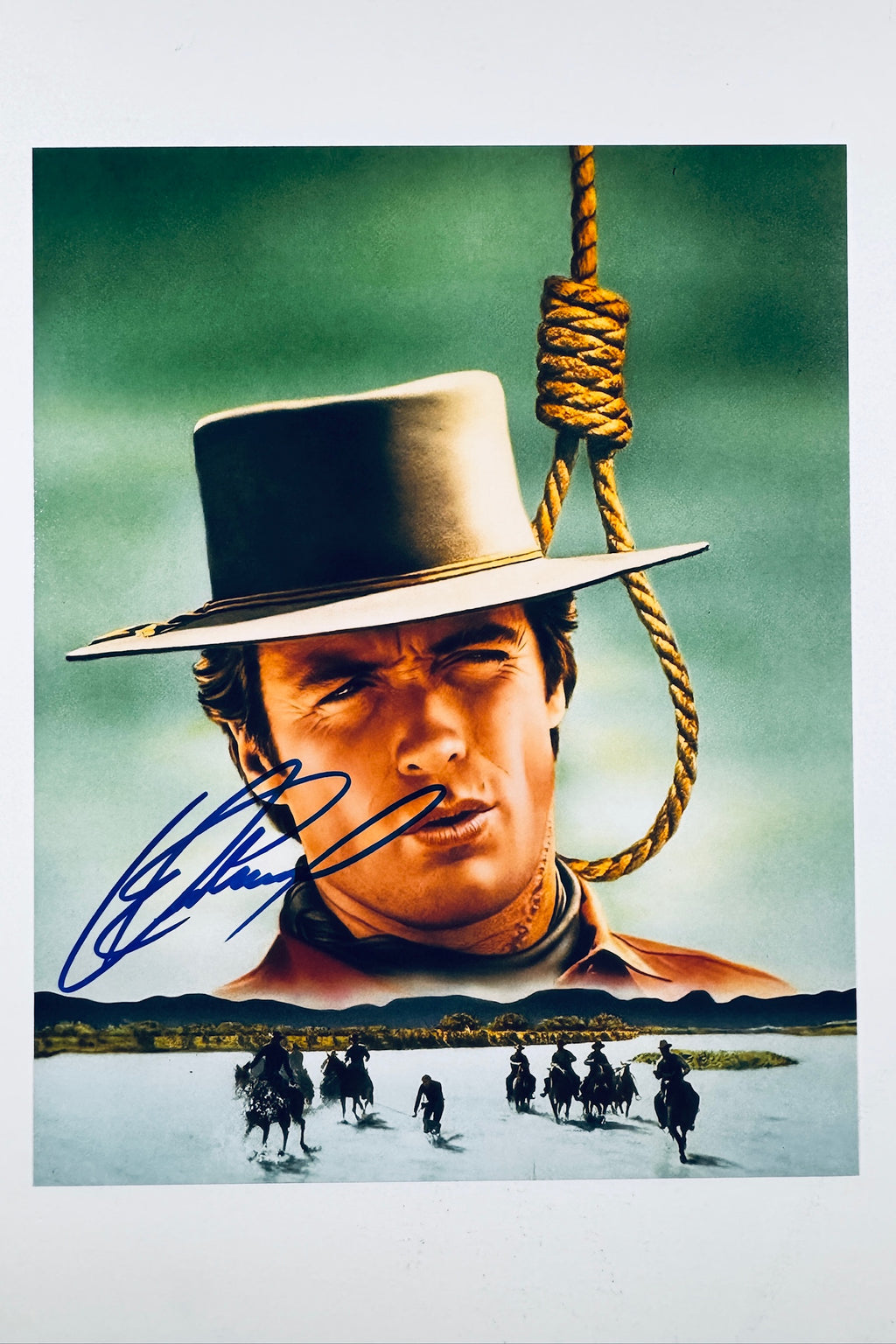 A promotional portrait of Clint Eastwood in a wide-brimmed hat beneath a hangman's noose with a landscape below, featuring a blue autograph signed across the lower left.