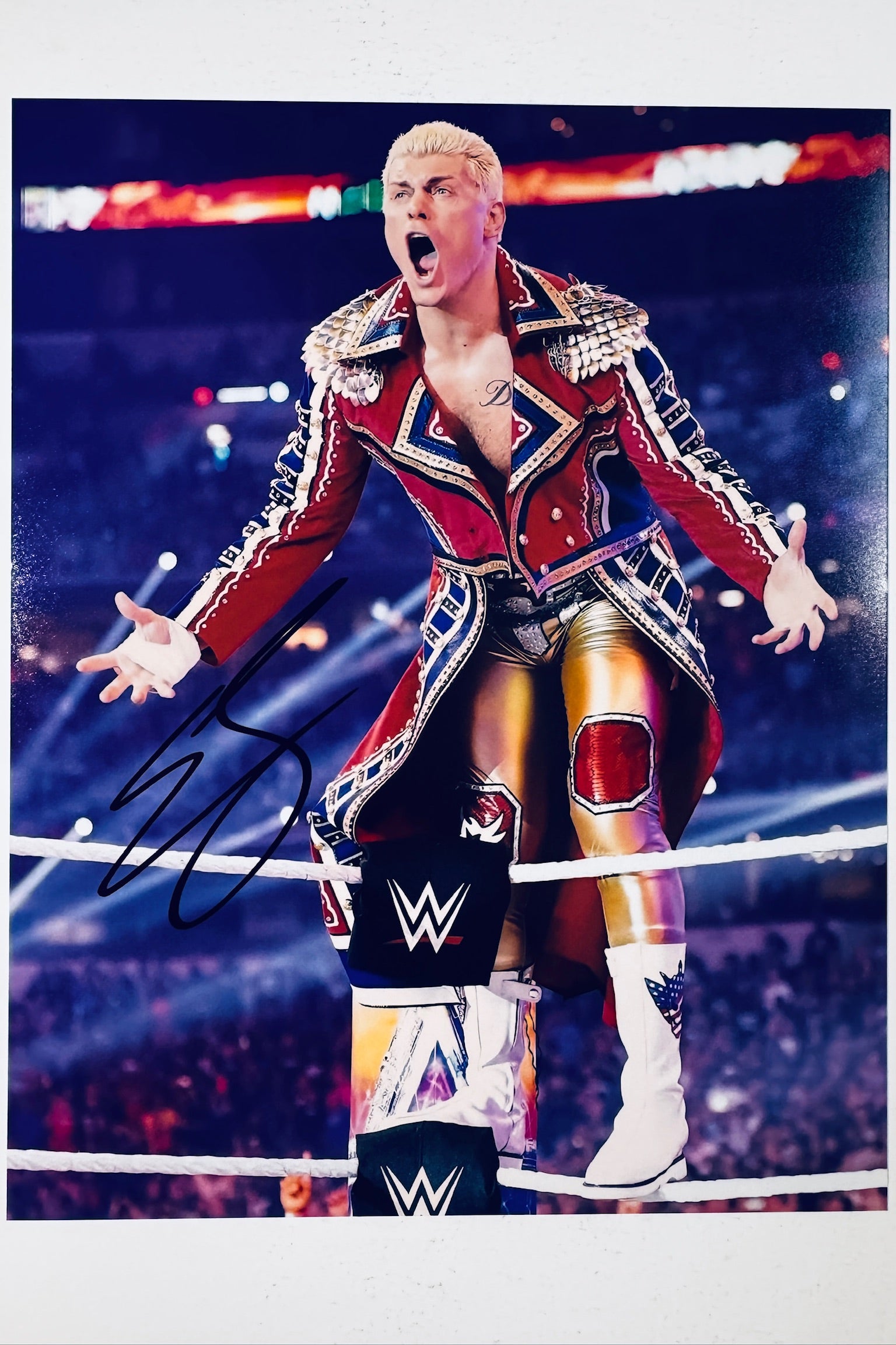 Signed full photo of Cody Rhodes standing on a ring turnbuckle in a red and blue ornate coat and gold wrestling gear, featuring a black autograph.