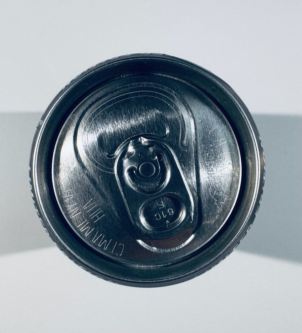Top-down view of a dark metallic beverage can lid with a pull-tab opening, photographed against a light gray background.