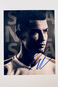 A close-up promotional portrait of Cristiano Ronaldo in a white jersey against a dark background with large lettering, featuring a blue marker signature across the lower right.