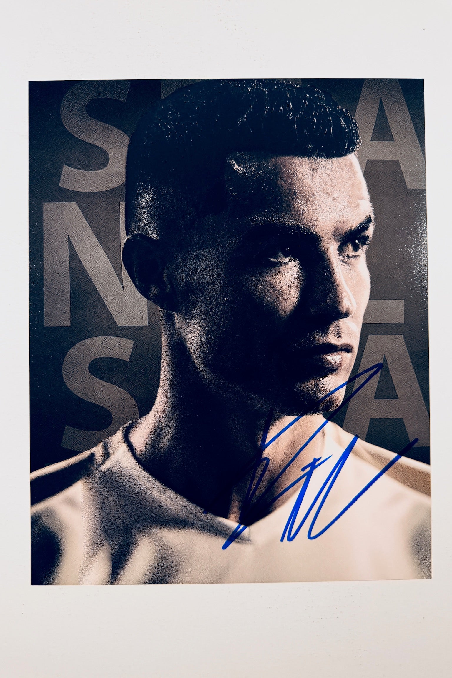 A close-up promotional portrait of Cristiano Ronaldo in a white jersey against a dark background with large lettering, featuring a blue marker signature across the lower right.