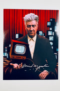 A portrait of filmmaker David Lynch sitting in a chair against a red curtain backdrop with several small vintage televisions stacked in the background while holding a small TV set in his hand, featuring a silver marker signature across the lower center.