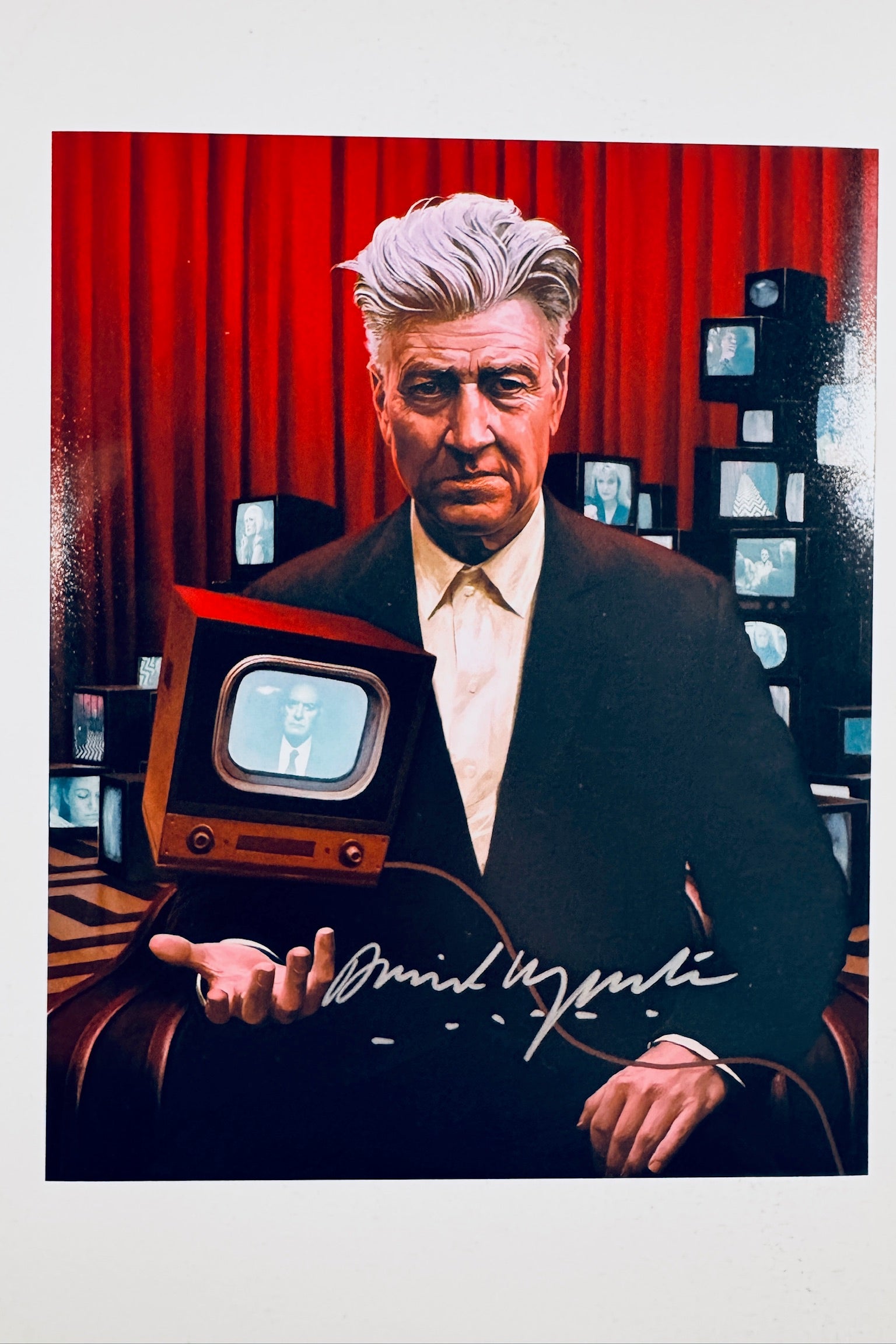 A portrait of filmmaker David Lynch sitting in a chair against a red curtain backdrop with several small vintage televisions stacked in the background while holding a small TV set in his hand, featuring a silver marker signature across the lower center.