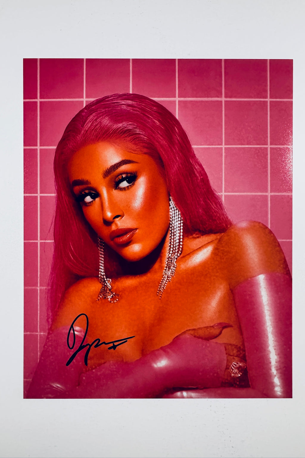 A portrait of Doja Cat with vibrant pink hair and long crystal earrings against a matching pink tiled background, featuring a black marker signature across the lower left.