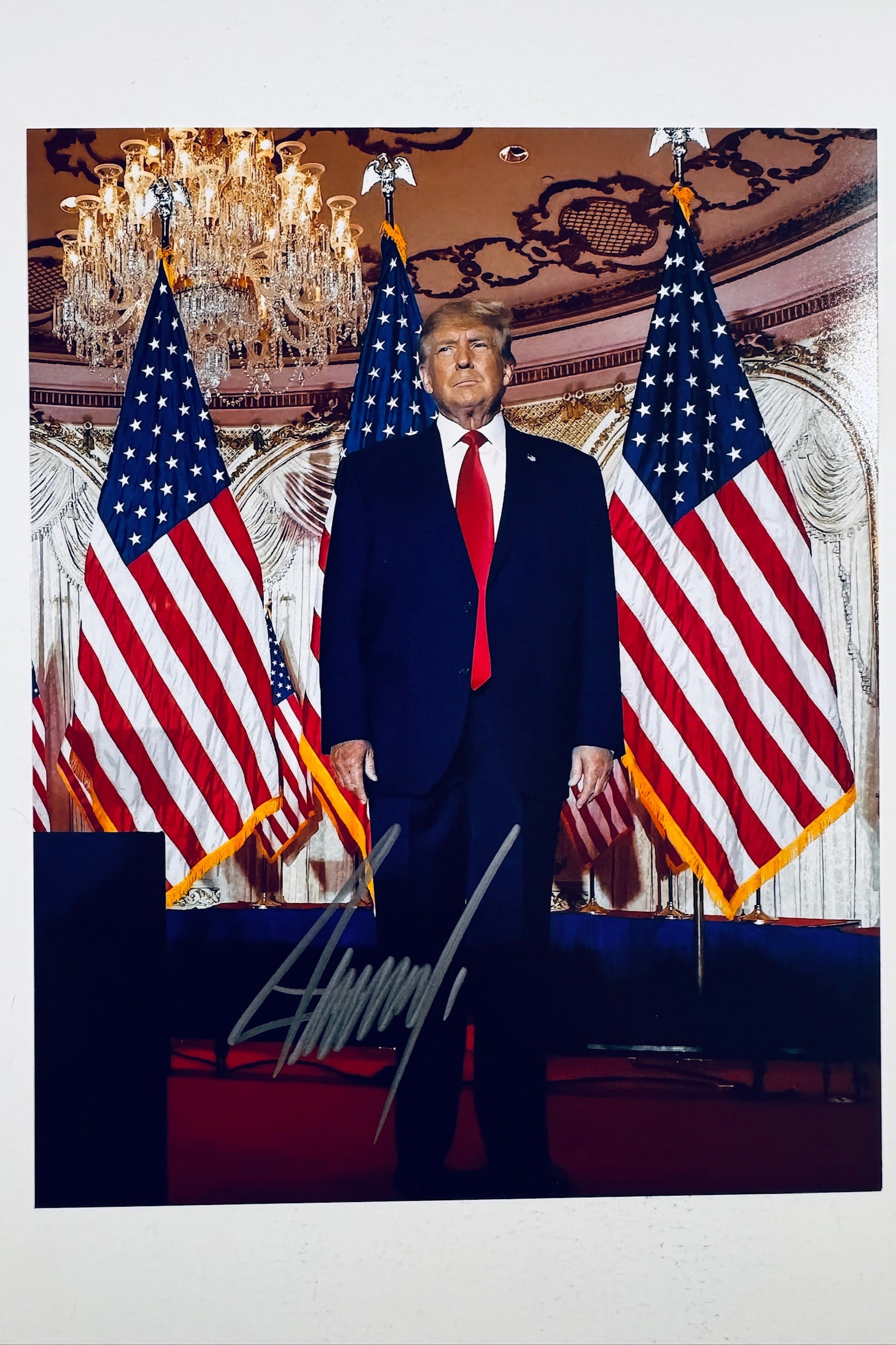 A portrait of Donald Trump standing in a formal room with American flags on either side, an ornate ceiling, and a crystal chandelier, featuring a dark marker signature across the lower center.