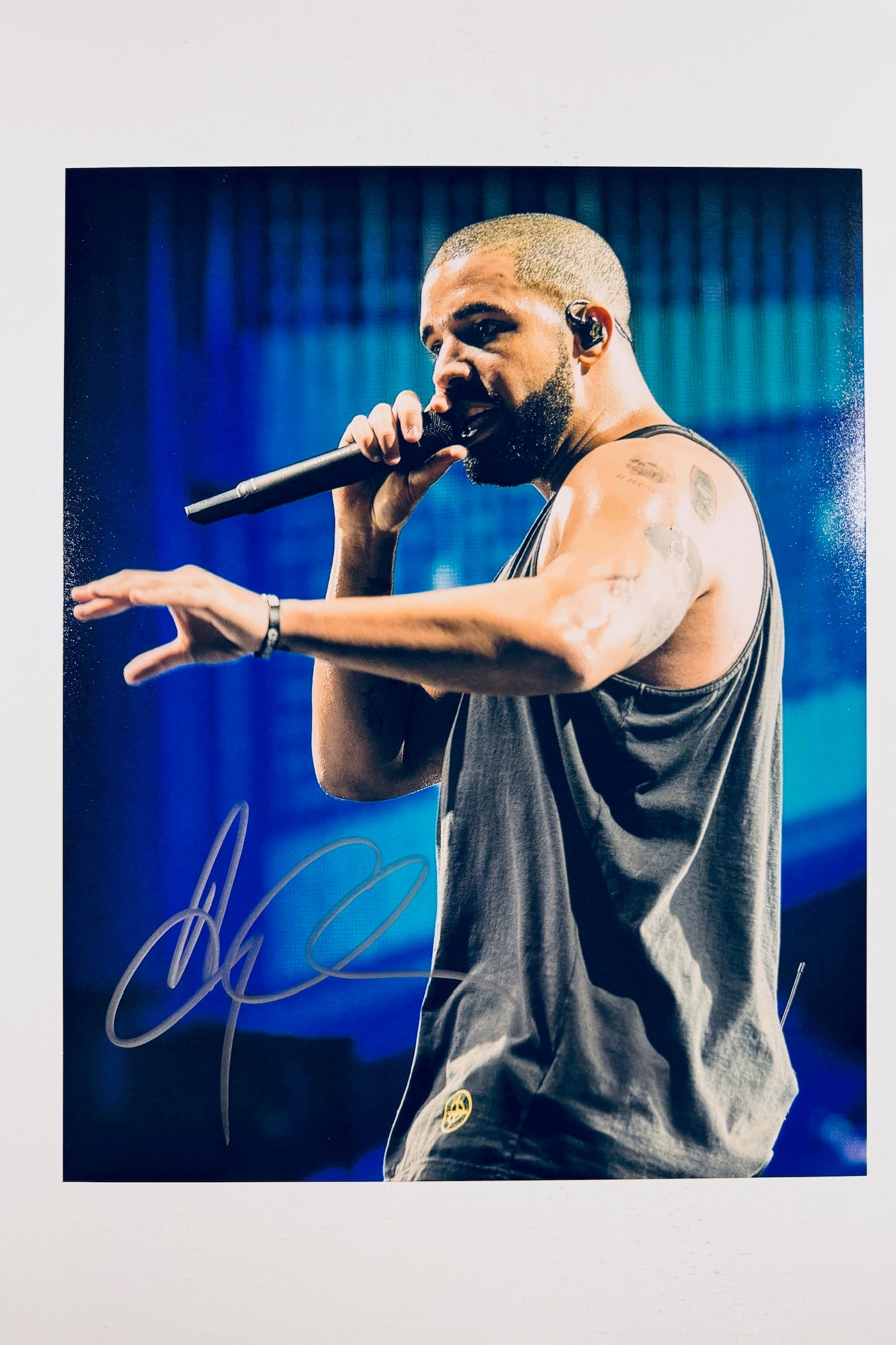 A profile portrait of Drake singing into a microphone on stage wearing a dark tank top against a blue background, featuring a silver marker signature across the lower left.