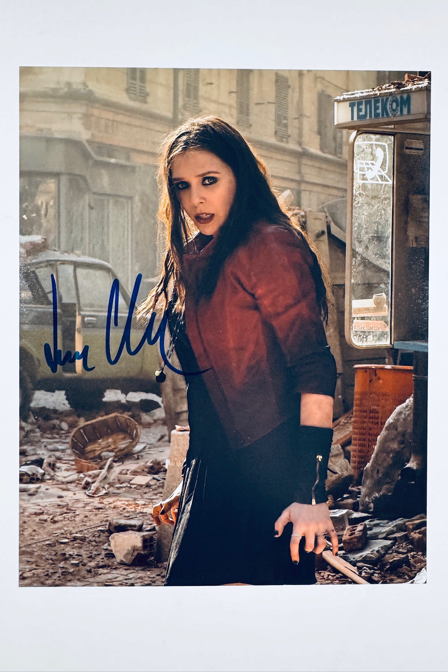 A still from Avengers featuring Elizabeth Olsen as the Scarlet Witch in a dark dress and red jacket amidst rubble with an intense look, signed in blue ink on the lower left.