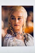 A close-up promotional portrait of Emilia Clarke as Daenerys Targaryen from Game of Thrones with braided blonde hair and a dragon-themed necklace, featuring a blue autograph across the lower portion.