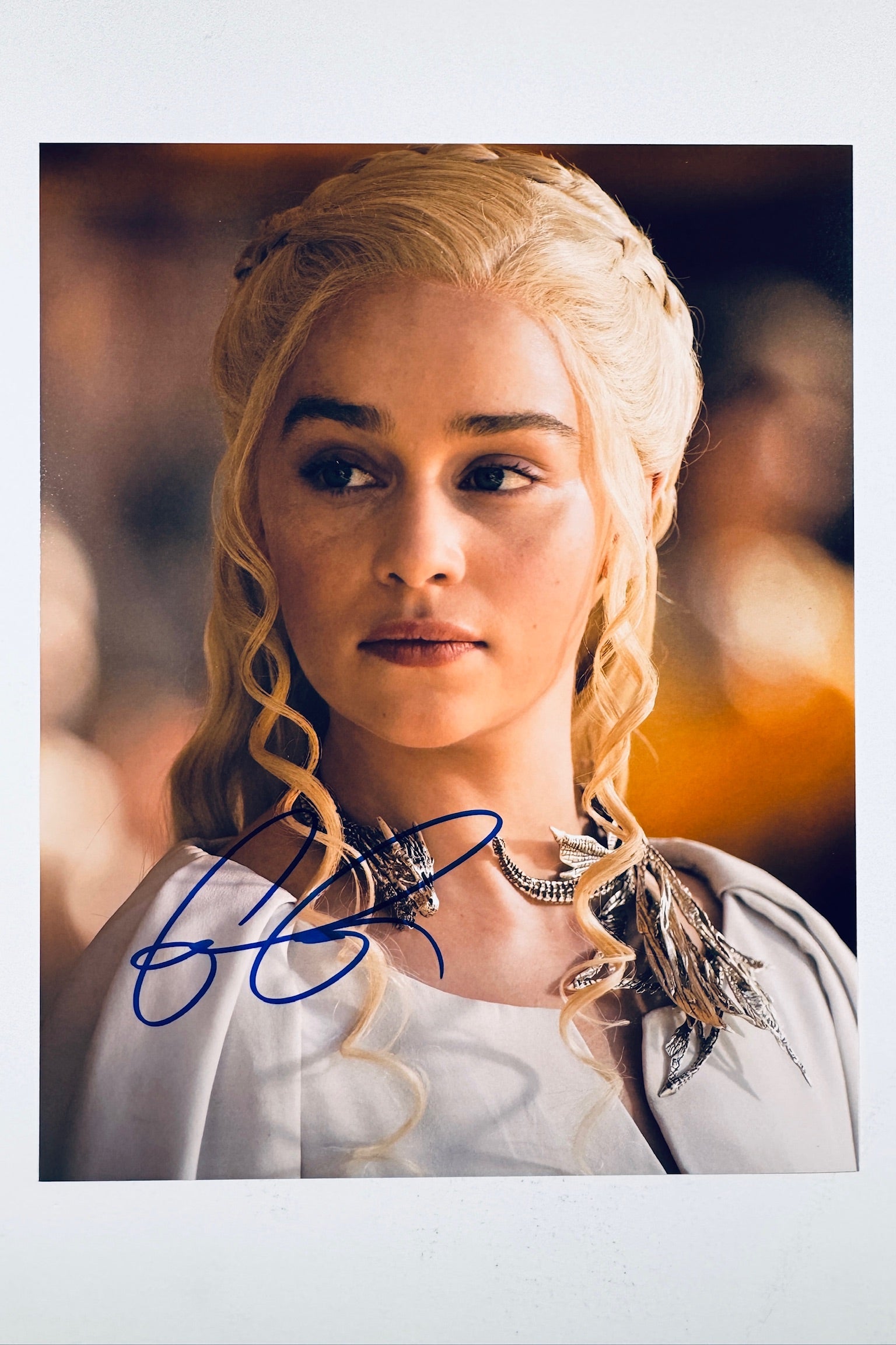 A close-up promotional portrait of Emilia Clarke as Daenerys Targaryen from Game of Thrones with braided blonde hair and a dragon-themed necklace, featuring a blue autograph across the lower portion.