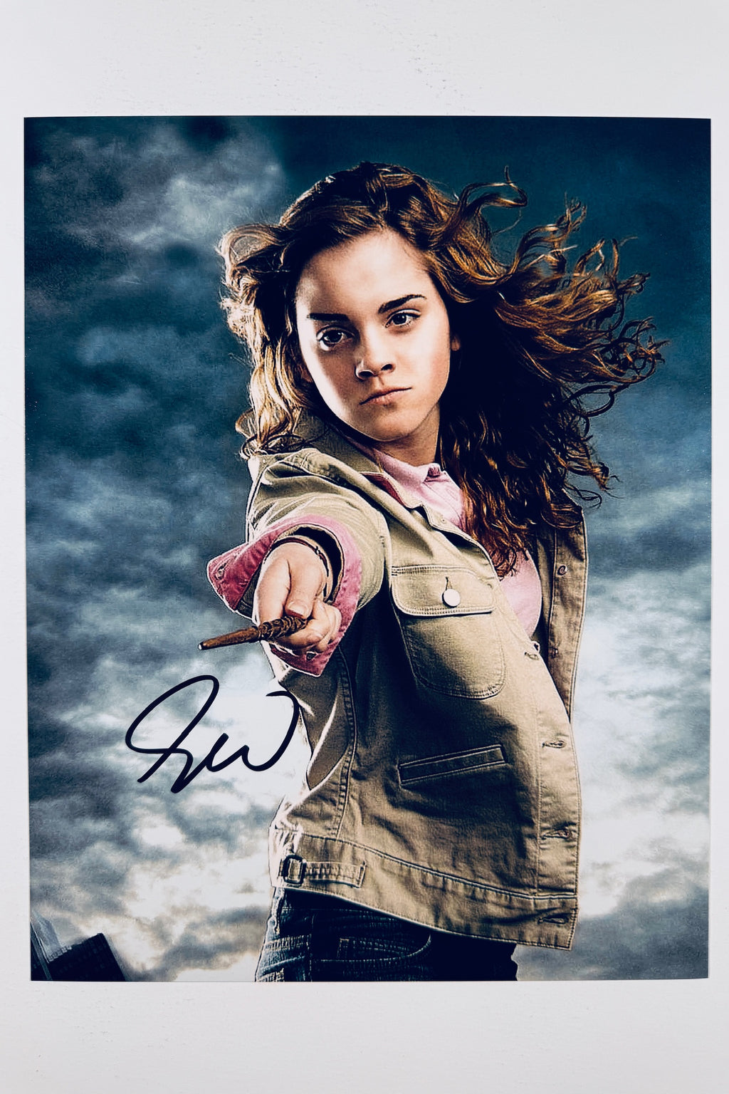 A still featuring Emma Watson as Hermione Granger pointing a wand forward under a cloudy sky, with a dark marker signature on the lower left.