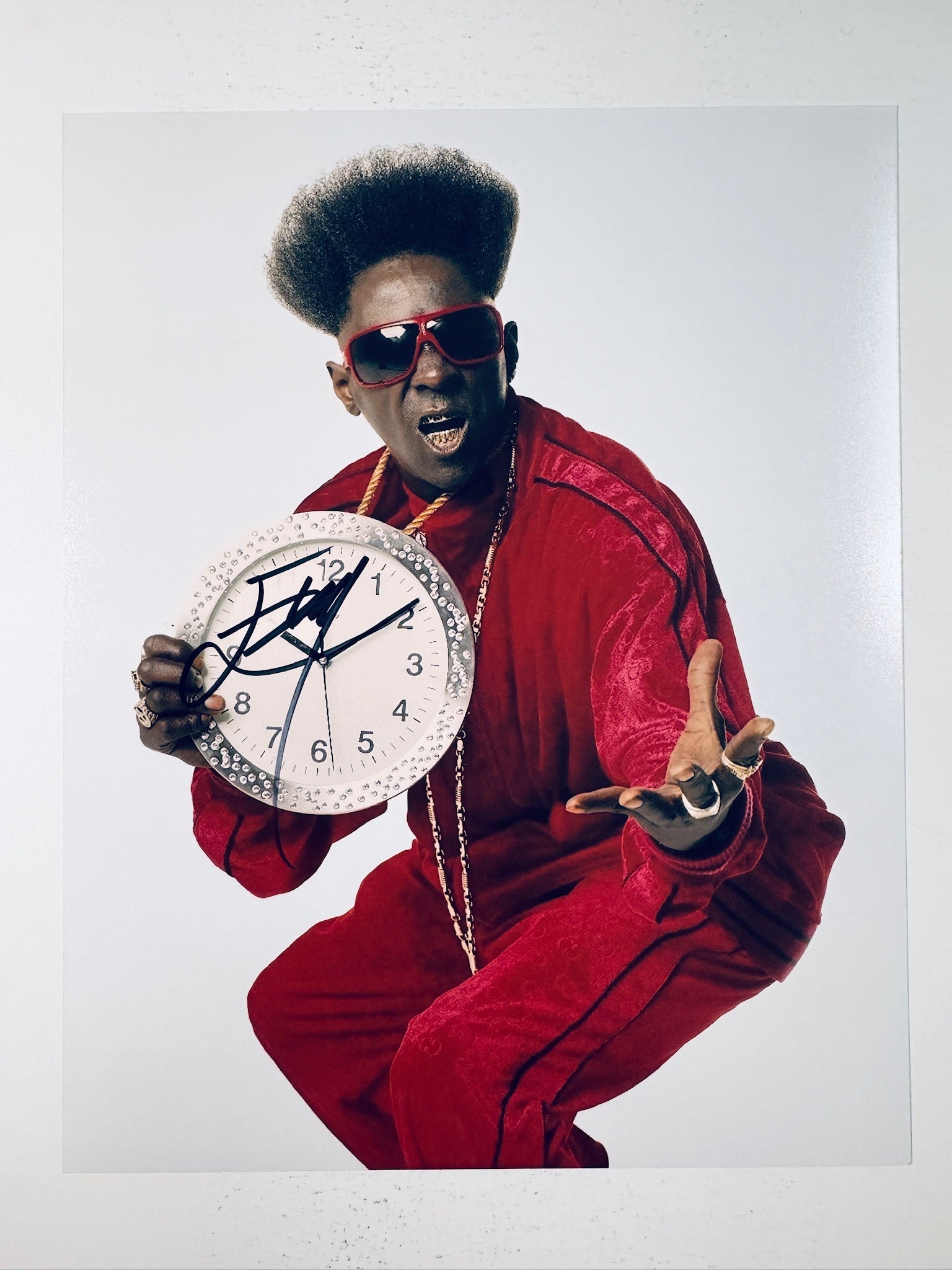 Signed 8x10 matte photo of Flavor Flav wearing a red velour outfit and red sunglasses while holding a large rhinestone clock, featuring a bold black ink autograph across the clock face.