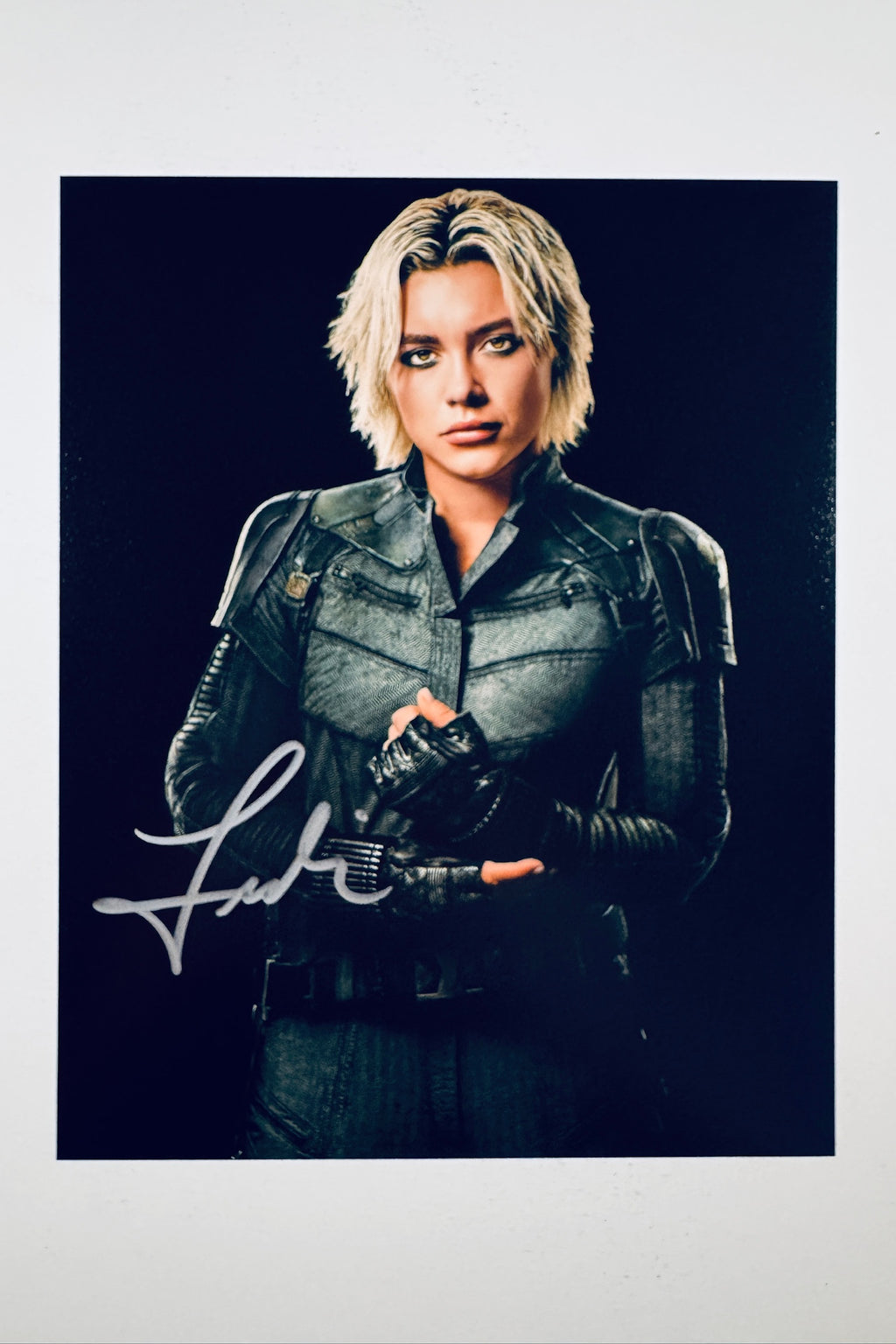 A promotional portrait of Florence Pugh as Yelena Belova with blonde hair and dark eye makeup, wearing a dark tactical suit and gloves against a black background, featuring a silver marker signature across her lower left.