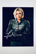 A promotional portrait of Florence Pugh as Yelena Belova with blonde hair and dark eye makeup, wearing a dark tactical suit and gloves against a black background, featuring a silver marker signature across her lower left.