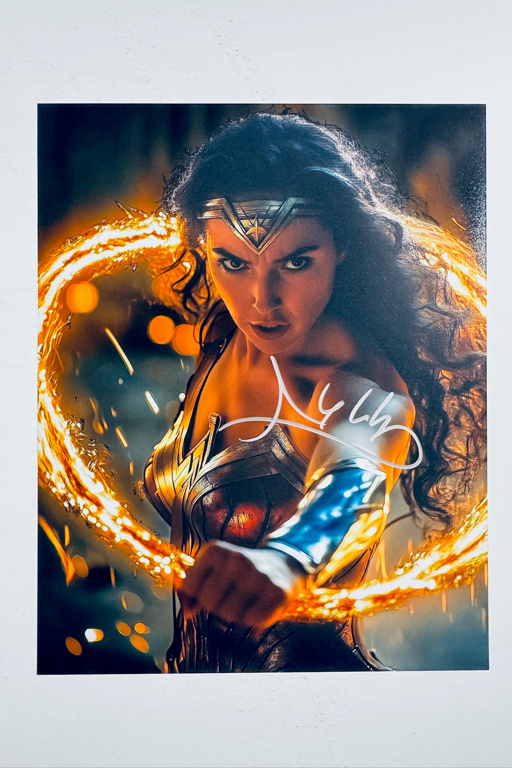 A close-up portrait of Gal Gadot as Wonder Woman wearing her tiara and armor while brandishing a glowing golden lasso of truth, featuring a silver marker signature across her midsection and arm.