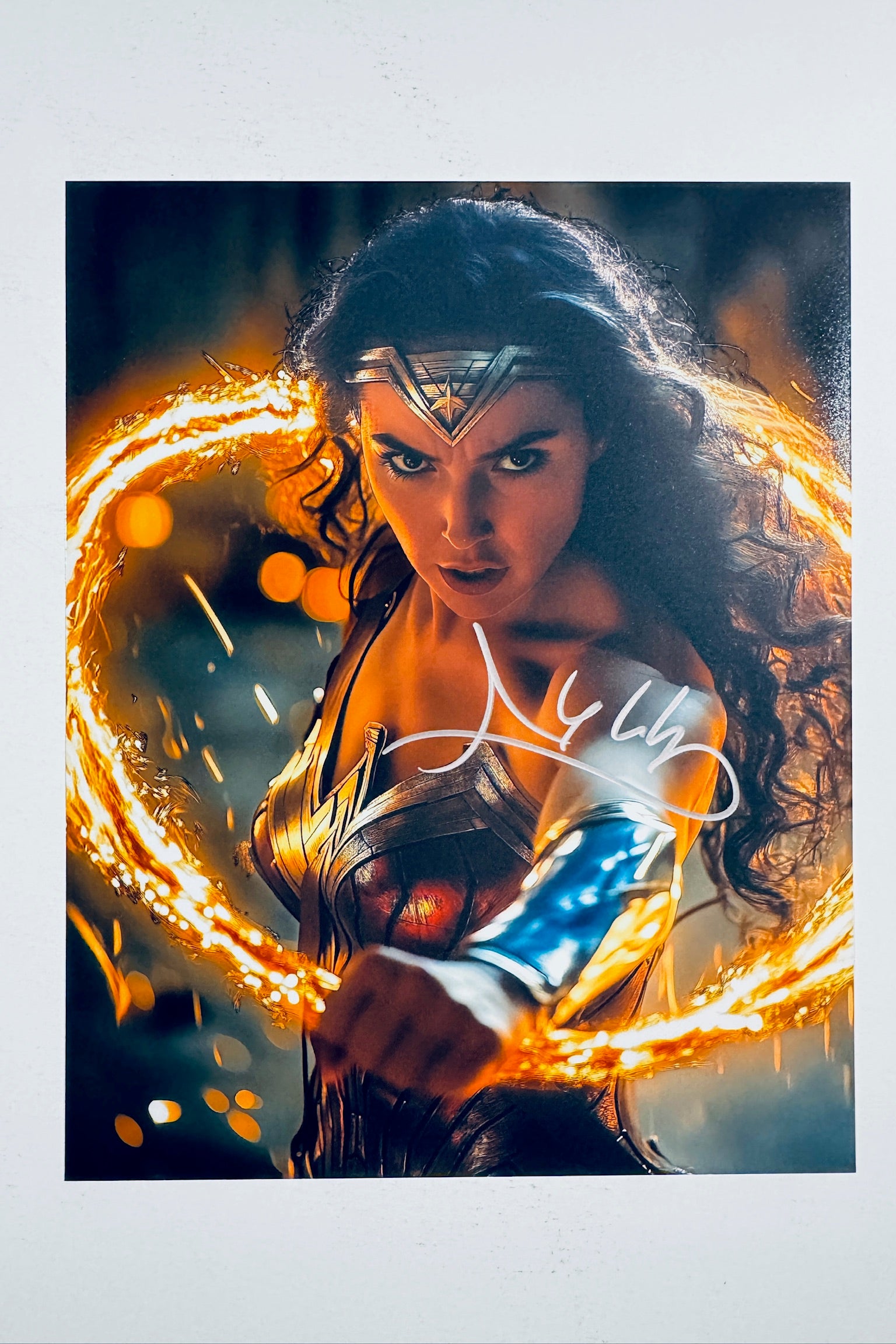 A close-up portrait of Gal Gadot as Wonder Woman wearing her tiara and armor while brandishing a glowing golden lasso of truth, featuring a silver marker signature across her midsection and arm.