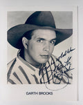 An autographed black and white portrait print of Garth Brooks wearing a cowboy hat, signed in blue ink with his name printed below on a white card against a light gray background.