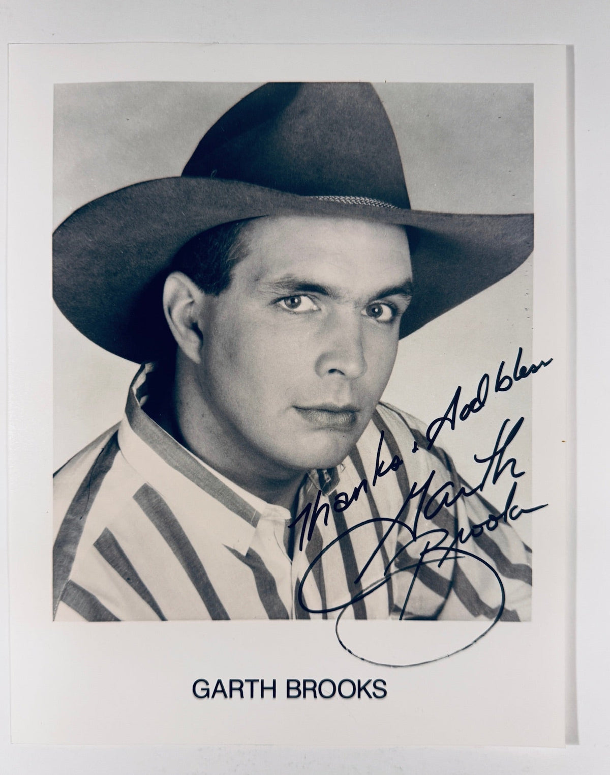 An autographed black and white portrait print of Garth Brooks wearing a cowboy hat, signed in blue ink with his name printed below on a white card against a light gray background.