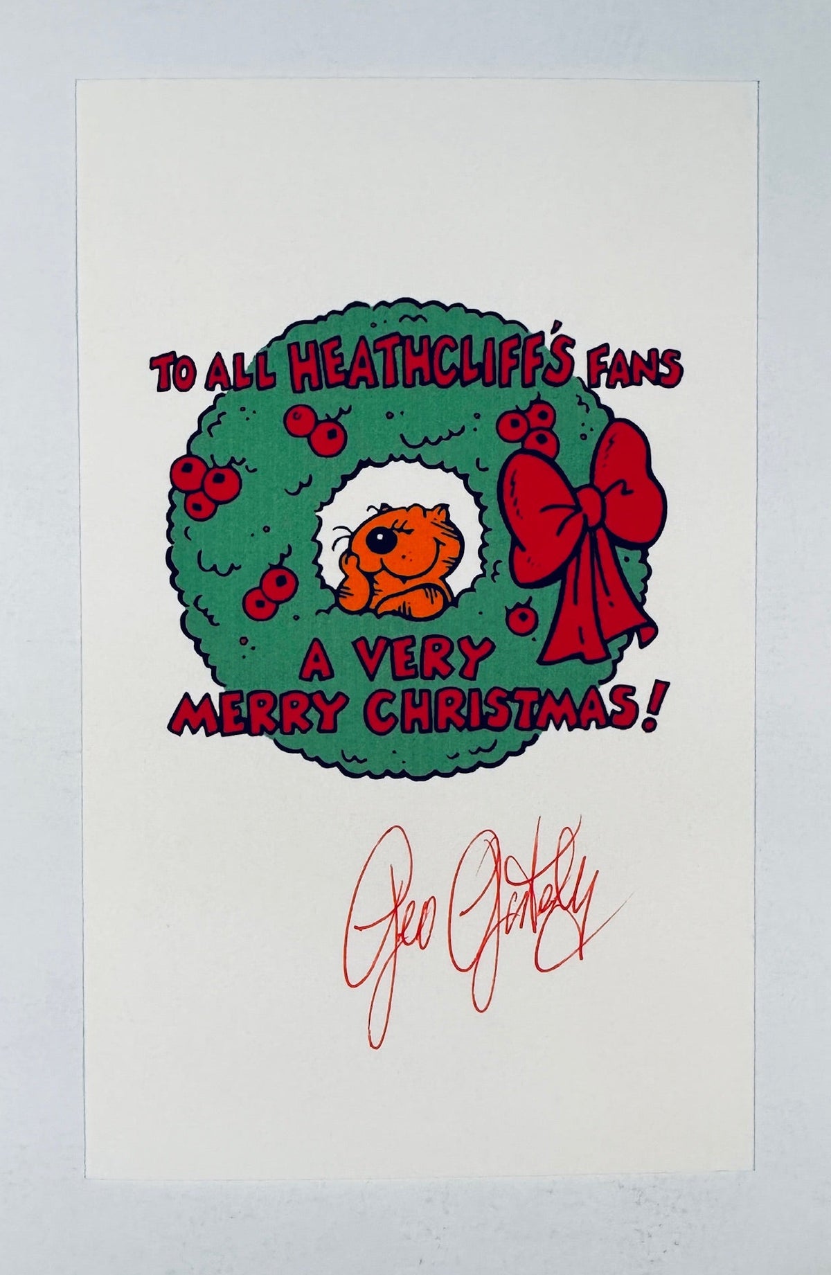 A white printed card featuring a Christmas wreath illustration with the text 'TO ALL HEATHCLIFF'S FANS A VERY MERRY CHRISTMAS!' and a red signature reading 'Geo Gently' below, photographed against a light gray background.