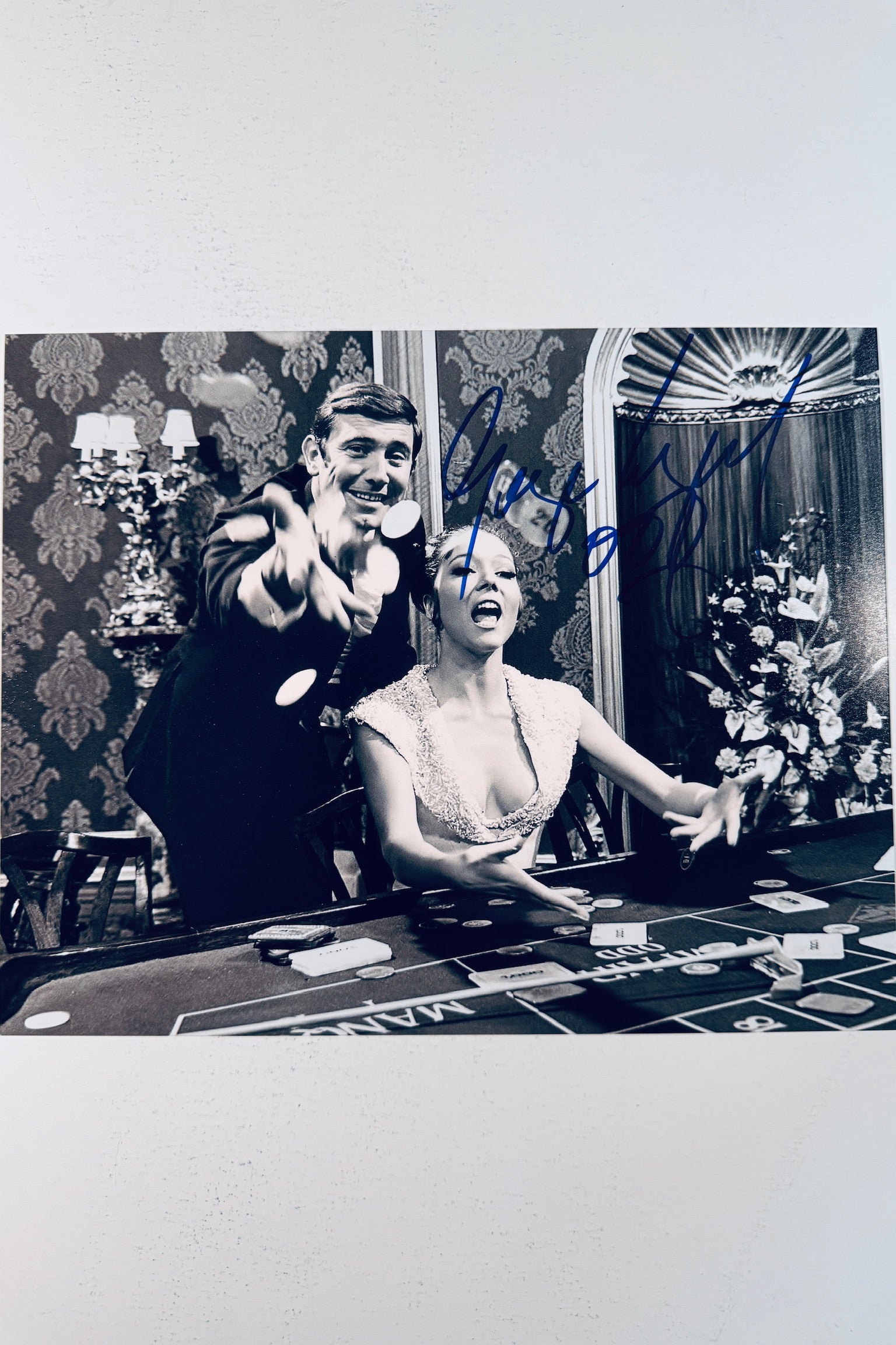 Signed 8x10 matte vintage black and white photo from On Her Majesty's Secret Service featuring George Lazenby and Diana Rigg at a casino table, with blue ink autographs by both actors.