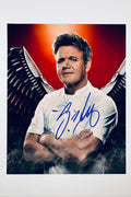 A promotional portrait of Gordon Ramsay in a white chef's coat with his arms crossed in front of a red background and dark, smoky angel wings, featuring a blue marker signature across his chest.